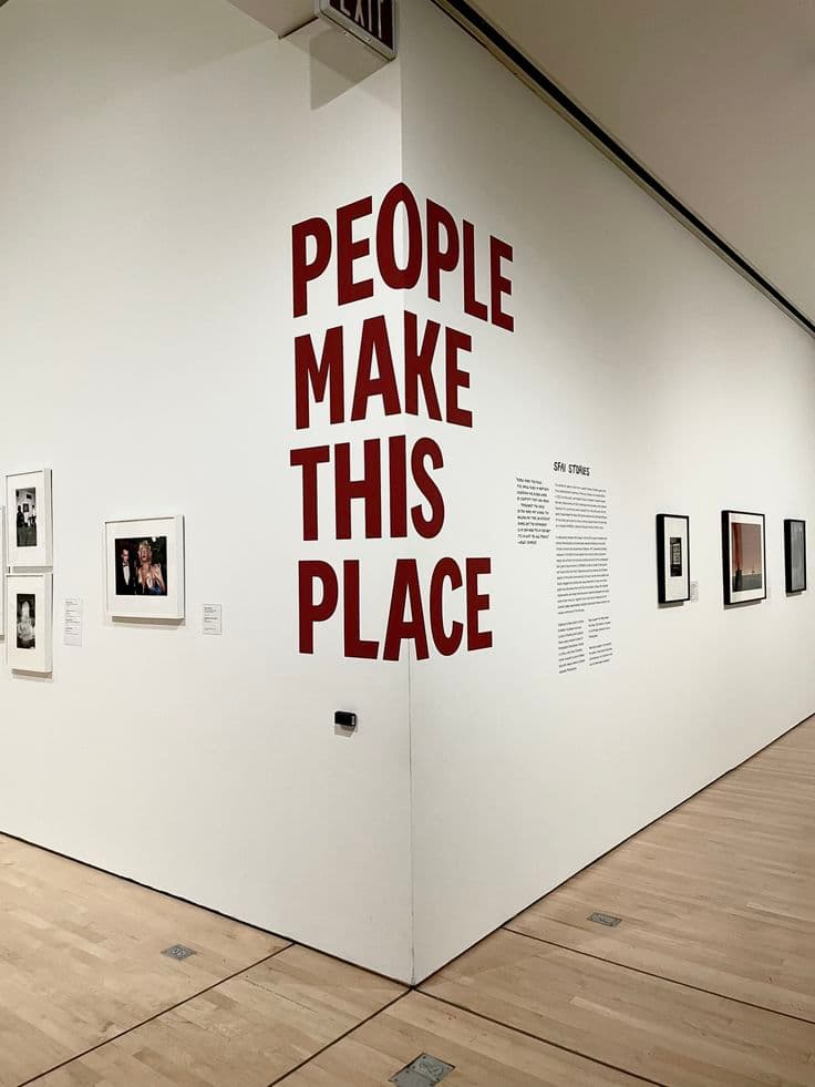 Texto 'PEOPLE MAKE THIS PLACE' em letras vermelhas grandes na quina de uma galeria branca.